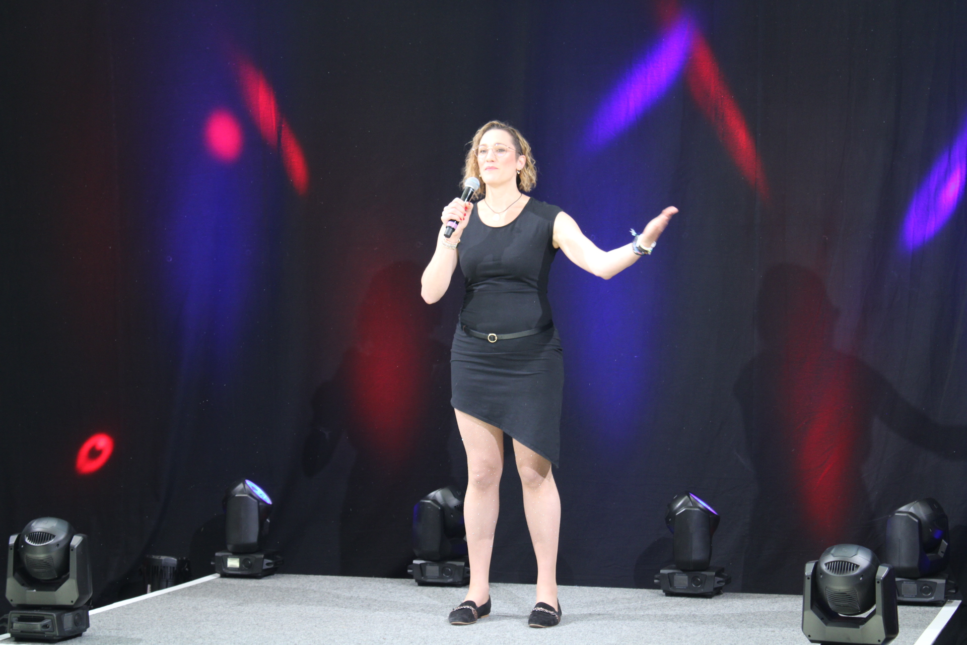Auf dem Bild ist in Ninja Abel zu sehen. Sie steht auf einer Bühne. Sie trägt ein schwarzes, kurzes Kleid mit Trägern. Sie spricht aktiv in ein Mikrofon, den linken Arm von sich auf schulterhöhe weggestreckt. Sie trägt ihre schulterlangen, blonden, leicht gewellten Haare offen, sie hat keinen Pony. Hinter ihr sieht man einen dunklen Hintergrund mit blauen und roten Lichtelementen. Die Bühne grau, auf der sie steht.